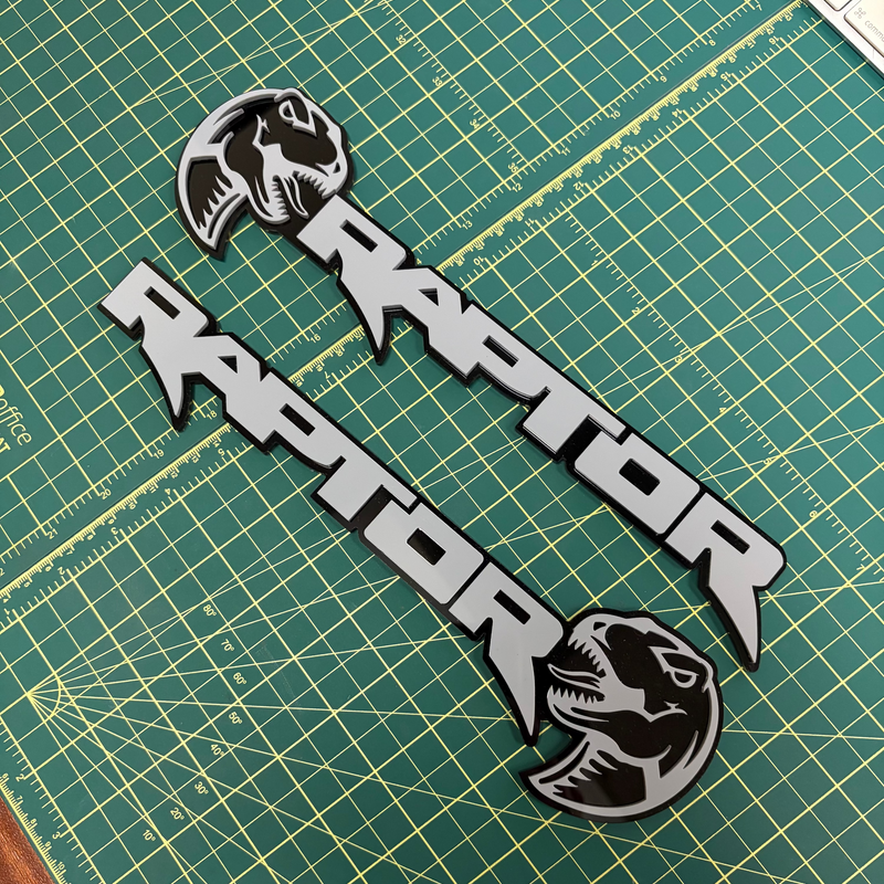 Load image into Gallery viewer, Two black and gray  'RAPTOR' emblems with dinosaur head graphics, displayed on a green cutting mat.
