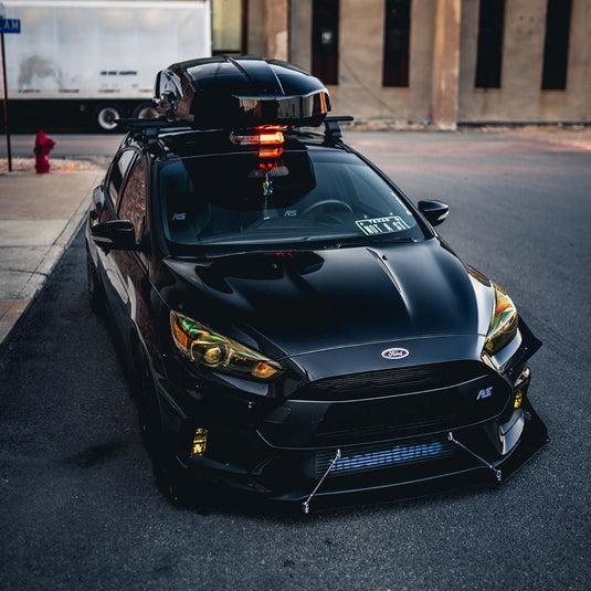 Ford Focus RS in Black with FSPE splitter support rods on the front end. Pictured on an asphalt street with a building and truck in the background. 